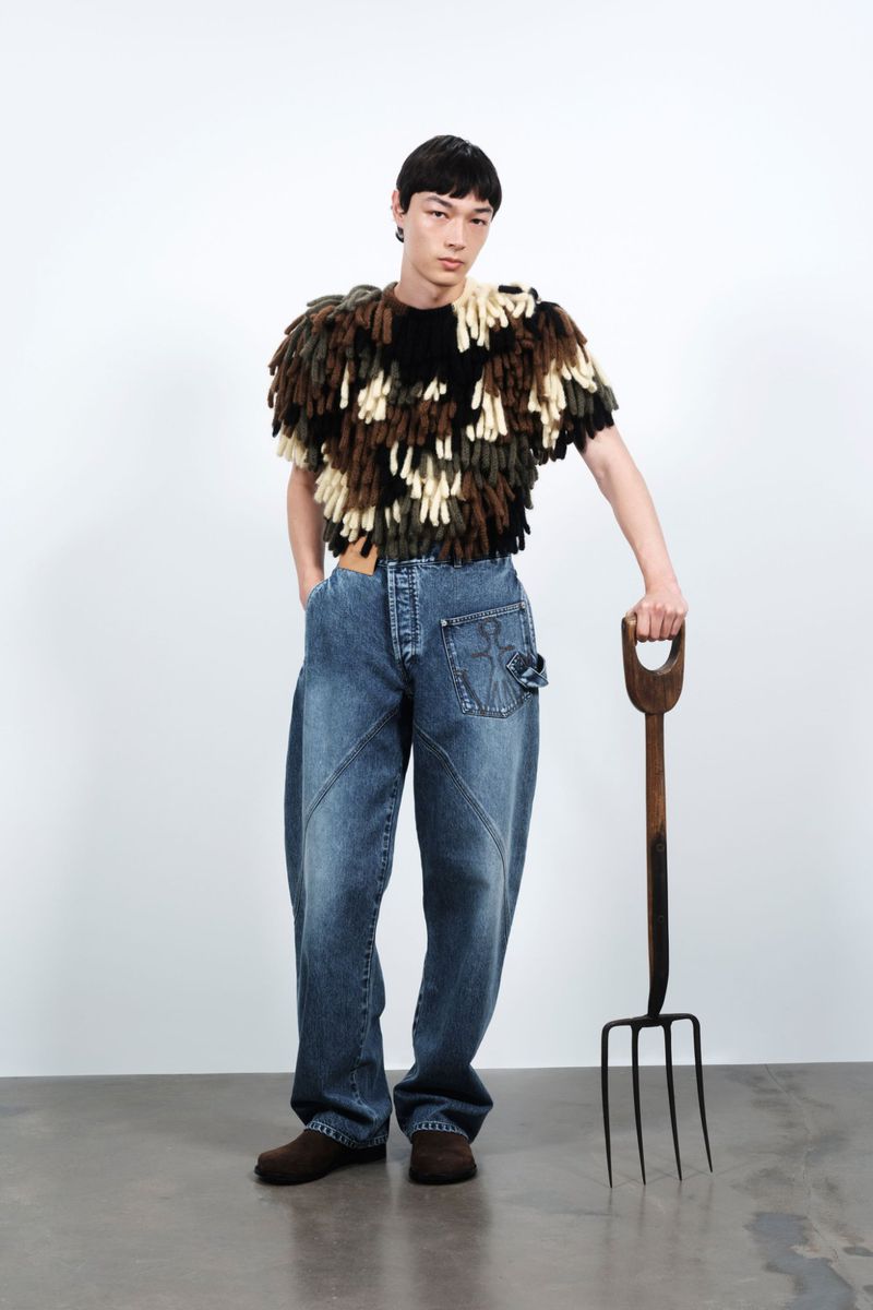 Asian male model posing in a fringed top and baggy denim pants, holding a metal pitchfork against a plain white backdrop in a studio shot.