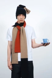 Model wearing a black beanie and multi-colored scarf holding a blue coffee cup against a plain white background