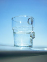 Silver chain rings draped over the rim of a clear glass of water under bright sunlight against a clear blue sky.
