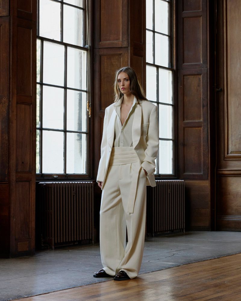 A woman poses in a beige suit in front of windows in an indoor setting, showcasing fashion and style with elegant and classic design.