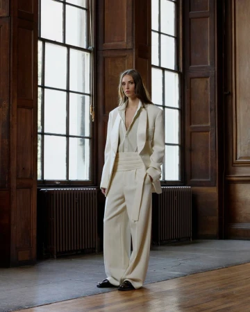 Model wearing a tailored off-white relaxed suit with wide-leg trousers standing in a wood-paneled room by tall windows.
