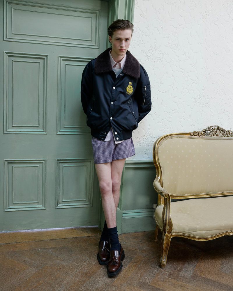 Young man in a bomber jacket poses in front of a paneled door, styled with dress shoes and socks next to an ornate cream chair.