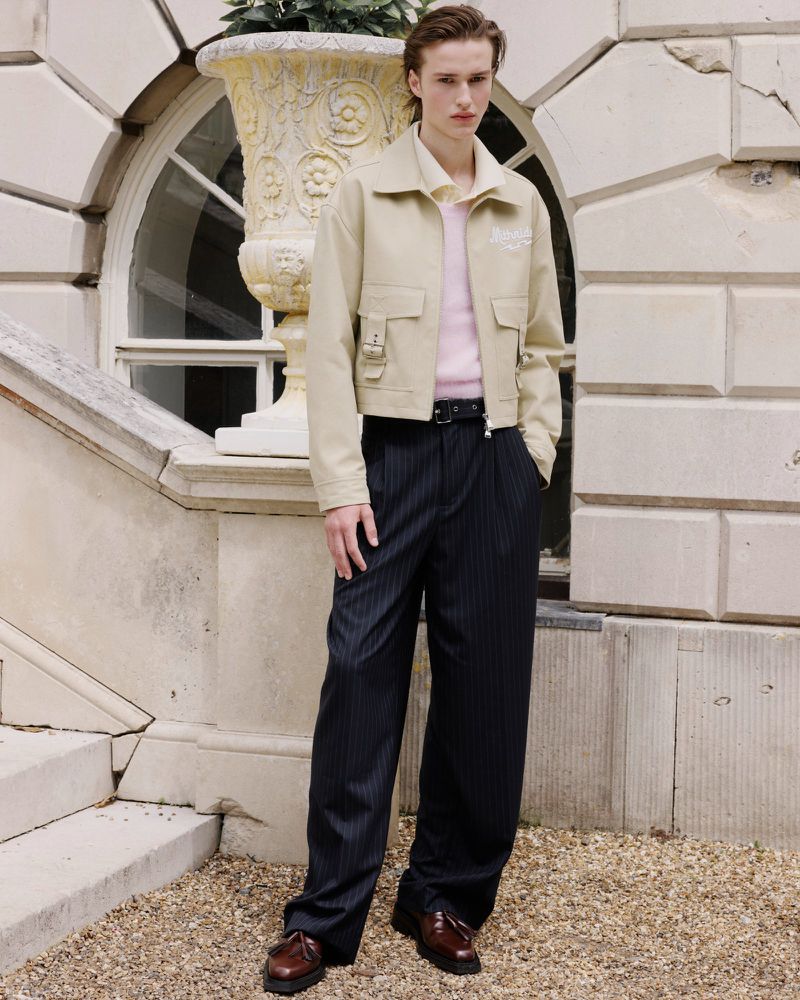 A fair-skinned adult male poses in a tan jacket and pinstripe pants beside a stone planter on a staircase in an outdoor setting.