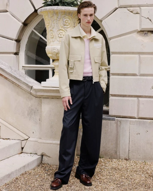 A fair-skinned adult male poses in a tan jacket and pinstripe pants beside a stone planter on a staircase in an outdoor setting.
