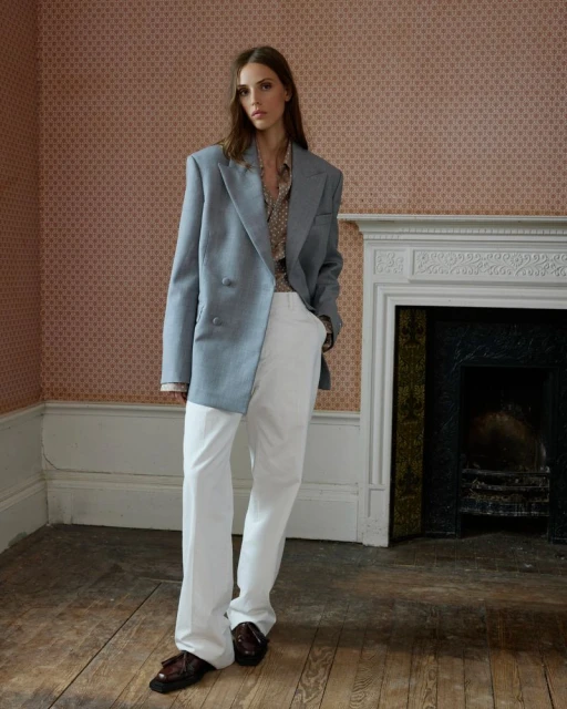 A fair-skinned young adult female is posing indoors in a light blue blazer, white pants, and loafers, standing in front of a fireplace.