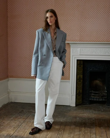 A fair-skinned young adult female is posing indoors in a light blue blazer, white pants, and loafers, standing in front of a fireplace.