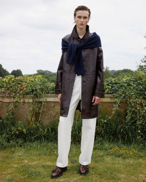 Full-shot of a young adult male fashion model in an outdoor setting, wearing a dark brown leather coat, white pants, and brown dress shoes.