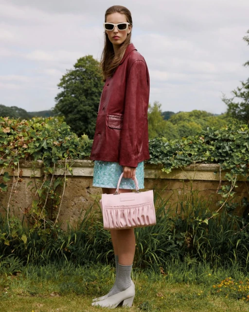 A woman stands on a grassy lawn, wearing a burgundy blazer, blue skirt, sunglasses, gray socks and heels, holding a pink purse.