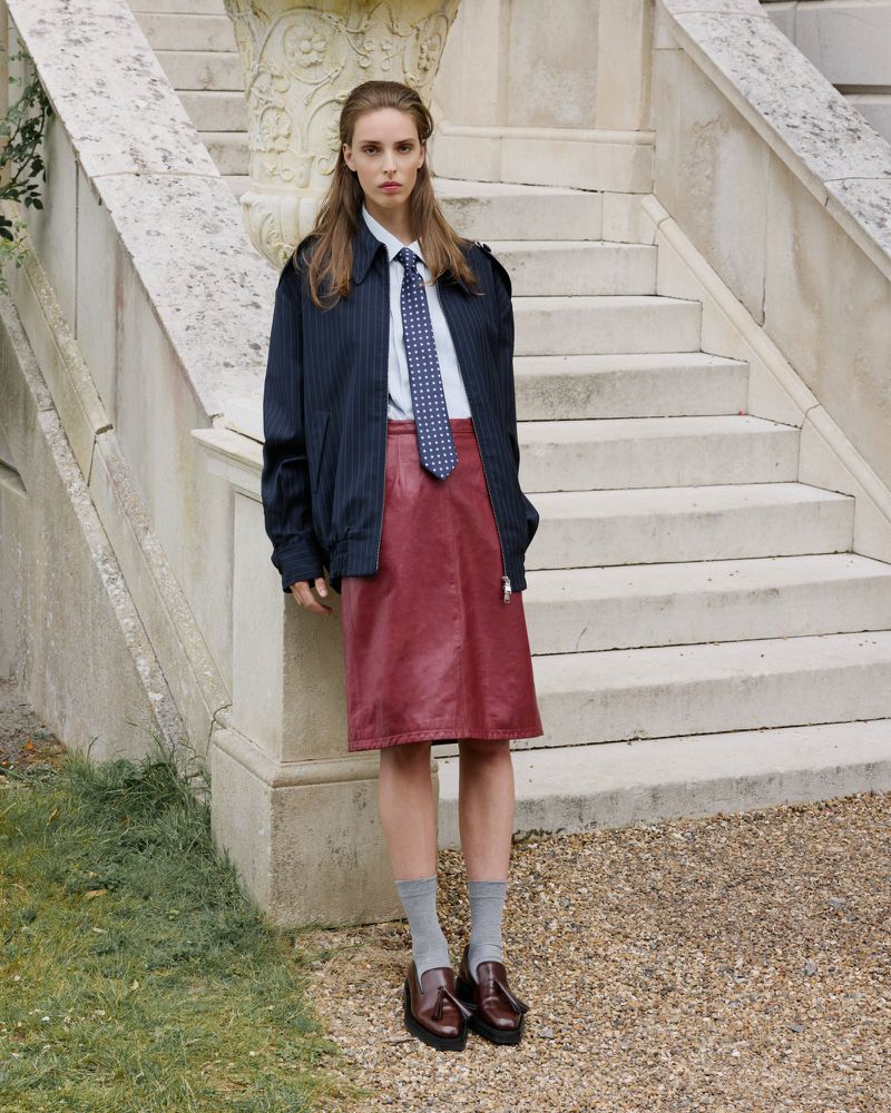 Woman in navy pinstripe jacket, collared shirt and tie, and maroon skirt stands on stone steps outdoors in a fashion editorial.