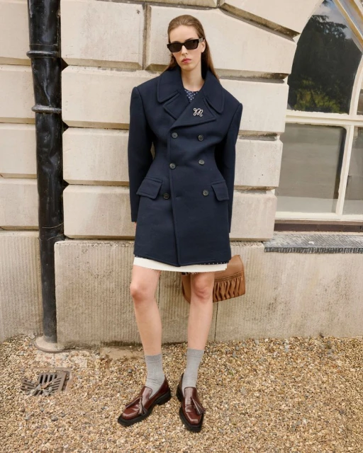 A woman poses in an oversized navy coat with a white dress, brown loafers, and a tan bag in front of a stone building.