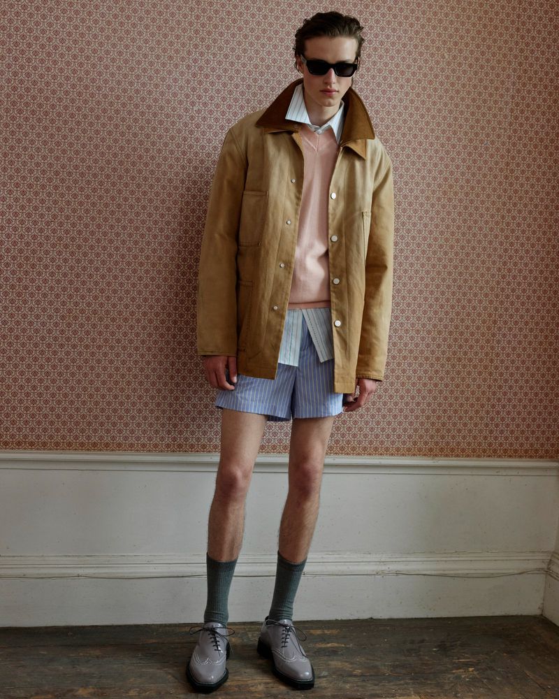 Man in tan jacket, light pink sweater, and blue shorts posing in front of patterned wallpaper, completing the fashionable ensemble.