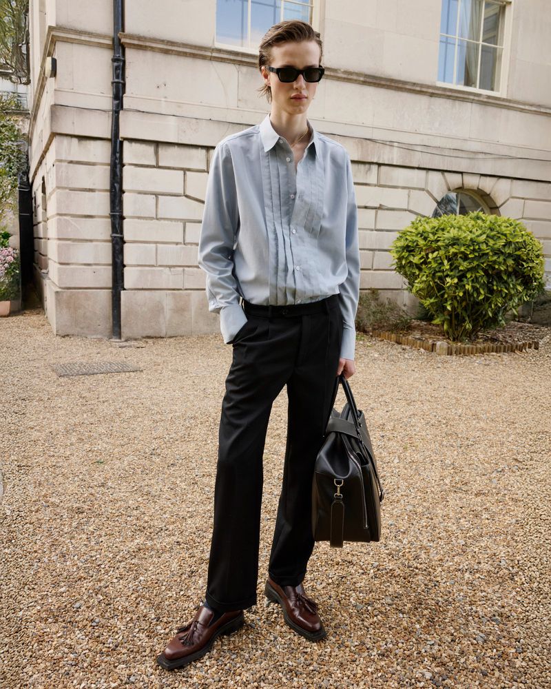 Male model in light blue button-down shirt, black pants, and sunglasses stands outdoors with a duffel bag, showcasing fashion.