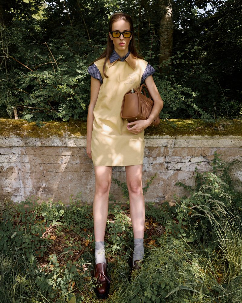 A woman in a short, pale-yellow dress stands holding a brown purse outdoors in front of a stone wall with a background of green foliage.
