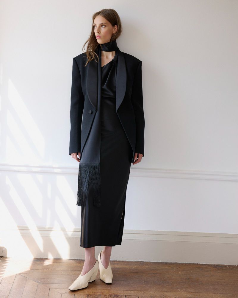 Model Martina Bergamini in a black tailored blazer, maxi dress with fringe scarf, and cream heels against a white interior wall.