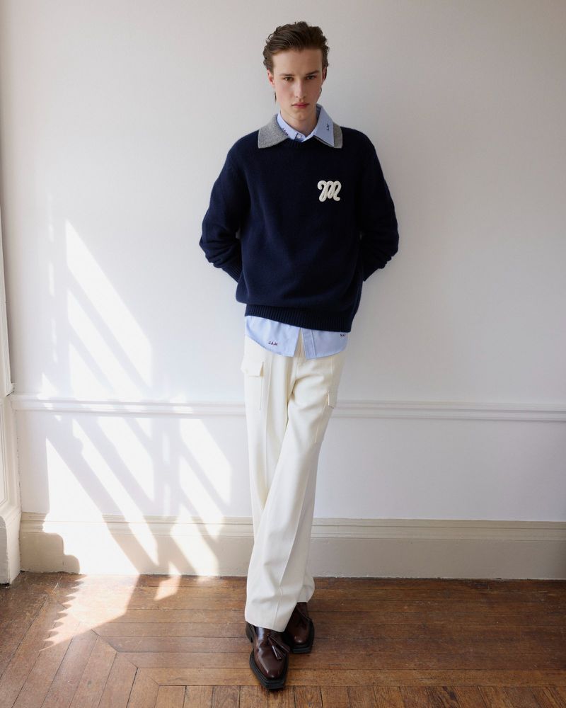 A stylish young man poses indoors wearing a navy blue crew neck sweater, light blue collared shirt, cream pants, and brown leather loafers.