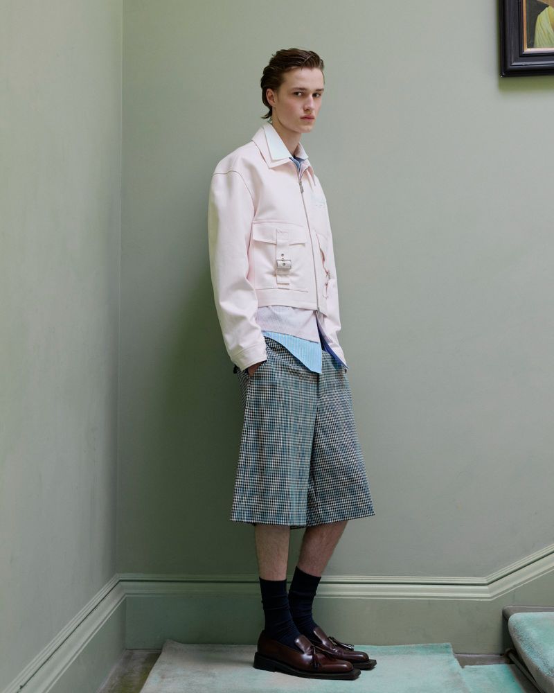 Fashionable male model poses on carpeted stairs, wearing a light pink jacket, patterned shorts, and brown leather shoes in a studio setting.