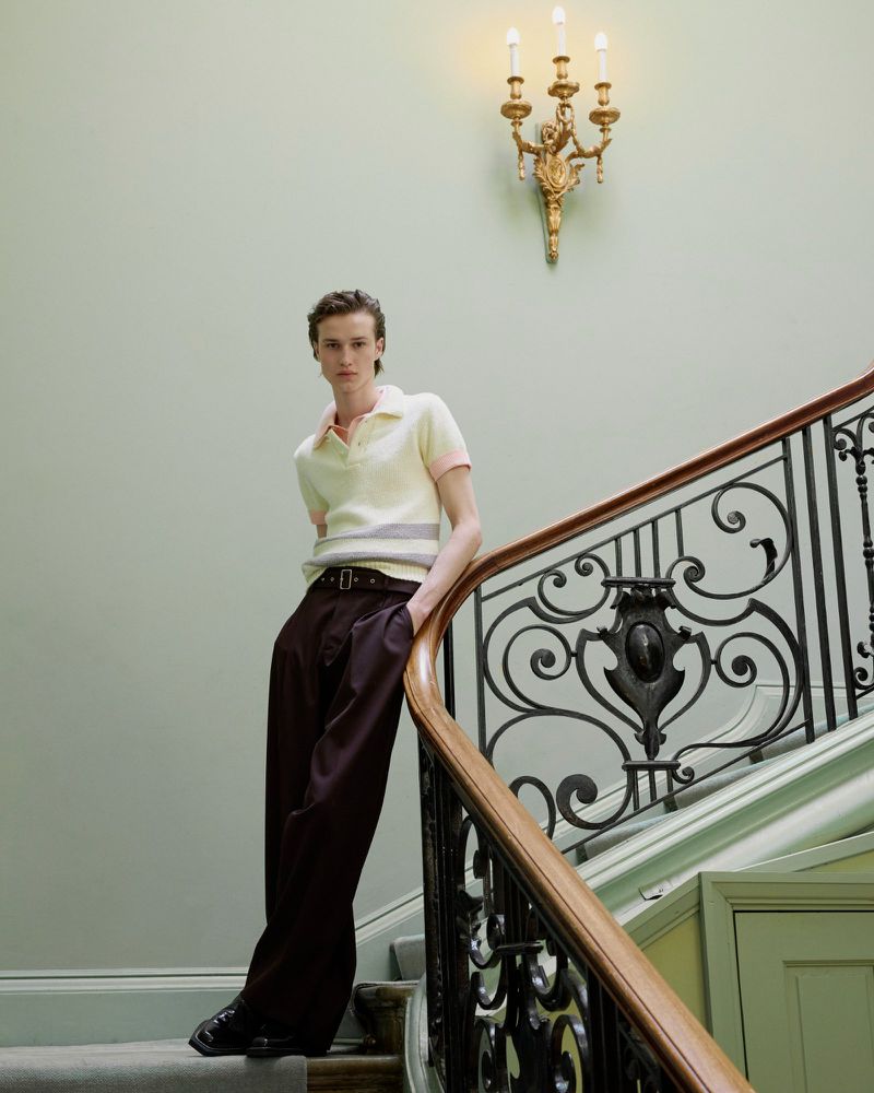 Indoor fashion shot of a slim adult male model on a staircase, wearing a yellow polo shirt and baggy brown pants.