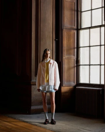A young adult woman stands in a light pink jacket and yellow shirt in a wood-paneled room with light from a window.