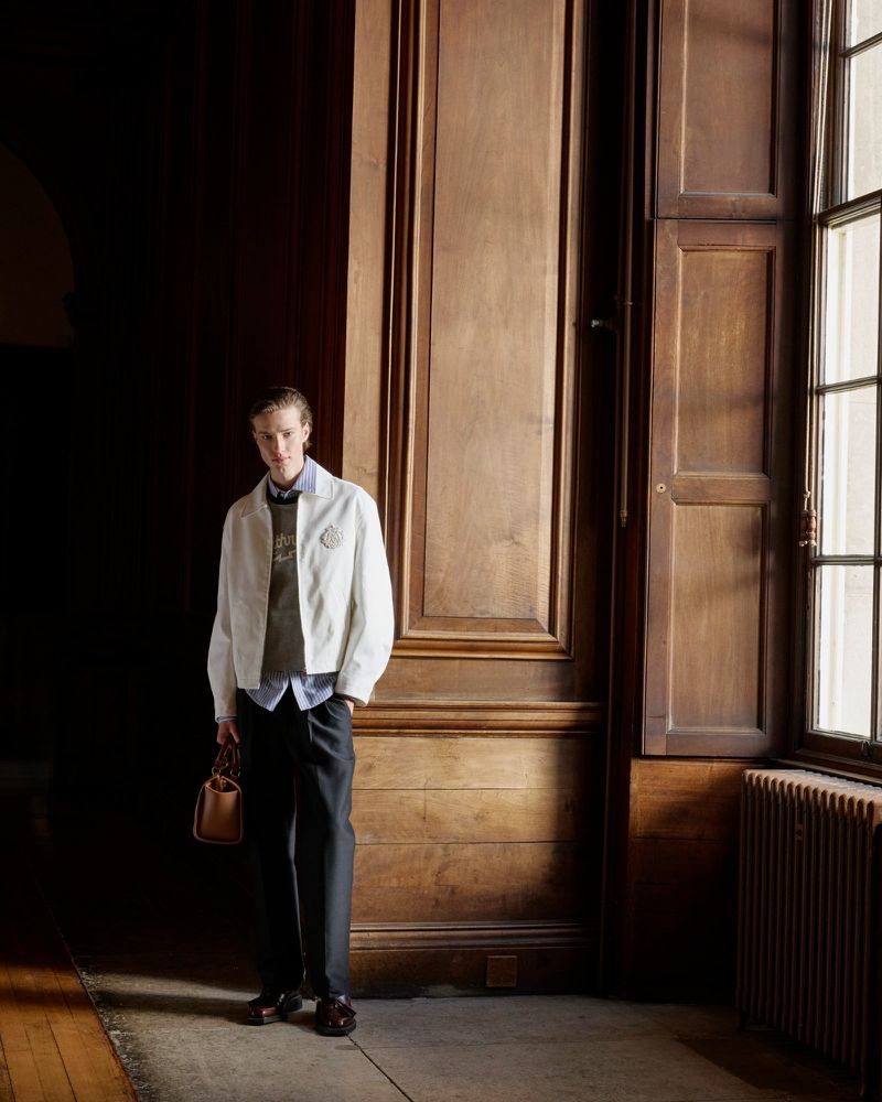 A blond male model poses indoors in a cream jacket, brown sweater, dark pants and brown loafers, holding a brown leather bag.