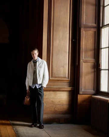 A blond male model poses indoors in a cream jacket, brown sweater, dark pants and brown loafers, holding a brown leather bag.