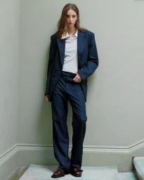 Model standing against a pale wall wearing a dark pinstripe blazer, matching trousers, and a white top for Mithridate.