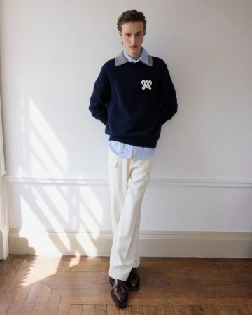 Model wearing a navy blue embroidered monogram sweater, layered collared shirt, and cream trousers leaning against a white wall.