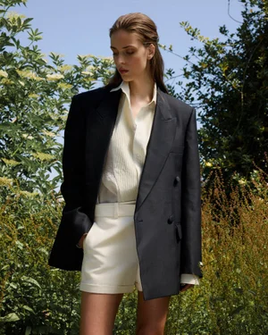 Model wearing an oversized dark blazer and cream shorts standing outdoors in a garden setting