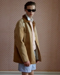 Model wearing a tan suede jacket, sunglasses, and striped shorts against patterned wallpaper