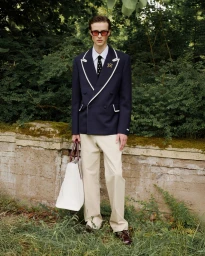 Model standing outdoors wearing a navy double-breasted blazer and holding a large tote bag in front of a low stone wall and foliage.