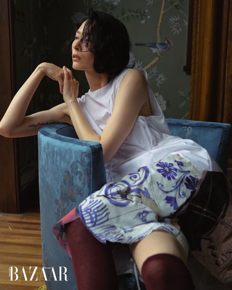Model Hsin-Yu Liu reclining in a blue velvet armchair wearing a white sleeveless top and printed skirt for Harper's Bazaar.