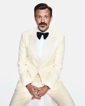 Actor Jason Sudeikis seated wearing an ivory tuxedo suit with a black bow tie against a white studio background.