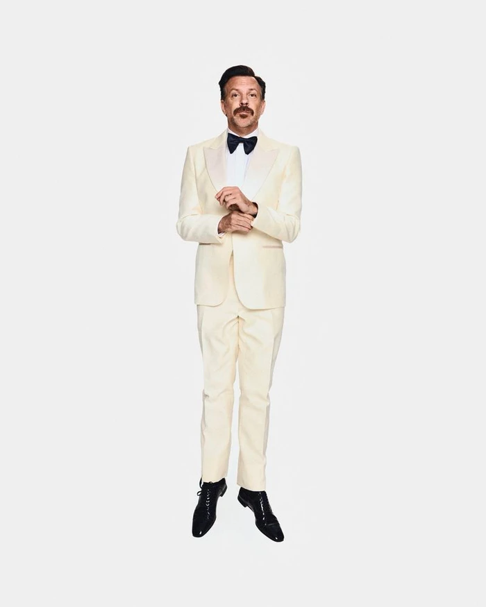 Jason Sudeikis suspended mid-air wearing a cream tuxedo and black bow tie against a pure white background.