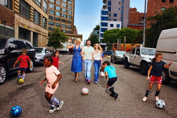 Hannah Waddingham, Jason Sudeikis, and Juno Temple walking on an urban street surrounded by kids playing soccer.