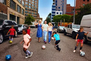 Hannah Waddingham, Jason Sudeikis, and Juno Temple walking on an urban street surrounded by kids playing soccer.