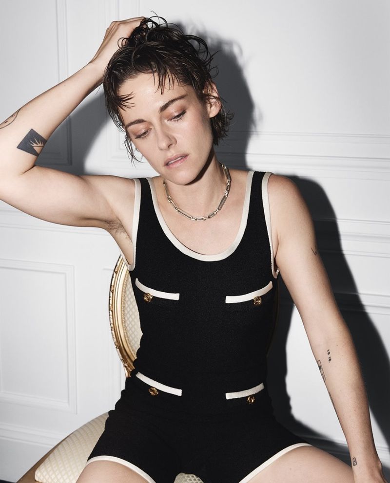 Kristen Stewart wearing a black knit romper with white trim while seated against a white paneled wall.