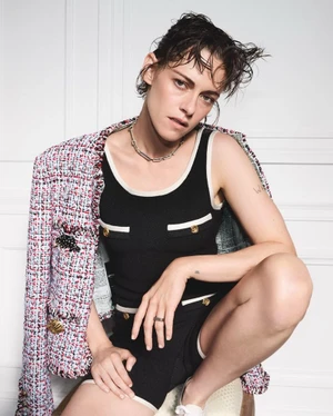 Kristen Stewart wearing a black knit romper with white trim and a draped tweed jacket against a white wall.