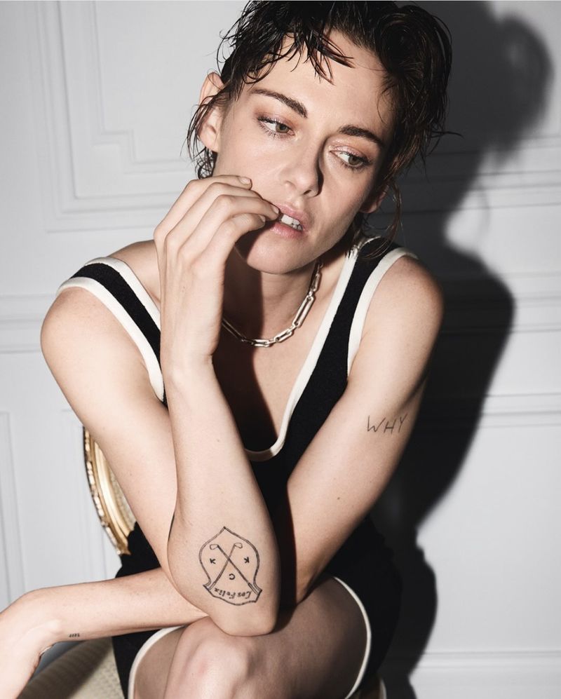 Kristen Stewart seated wearing a black tank dress with white trim, chain necklace, and short wet-look hair against a white wall.
