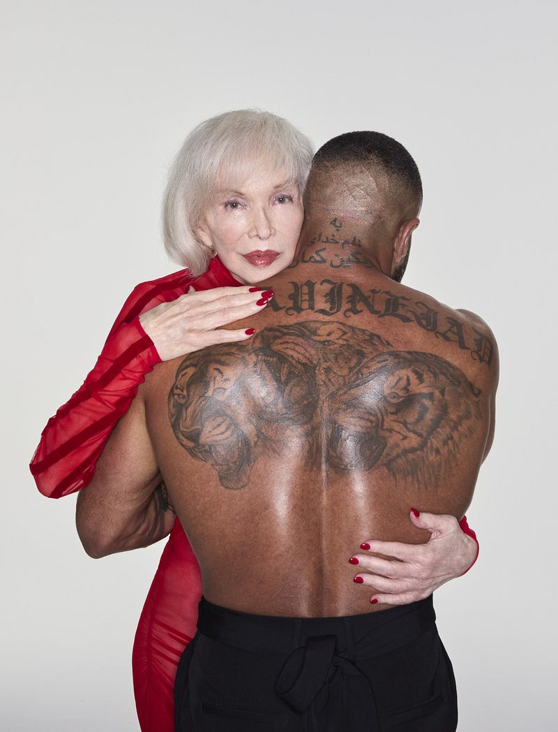 An elderly white woman in red embraces a Black man with tiger tattoos on his back against a white background in a studio setting.