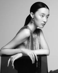 Black and white fashion portrait of a woman in a metallic chainmail top and hoop earrings resting her arms on a chair.