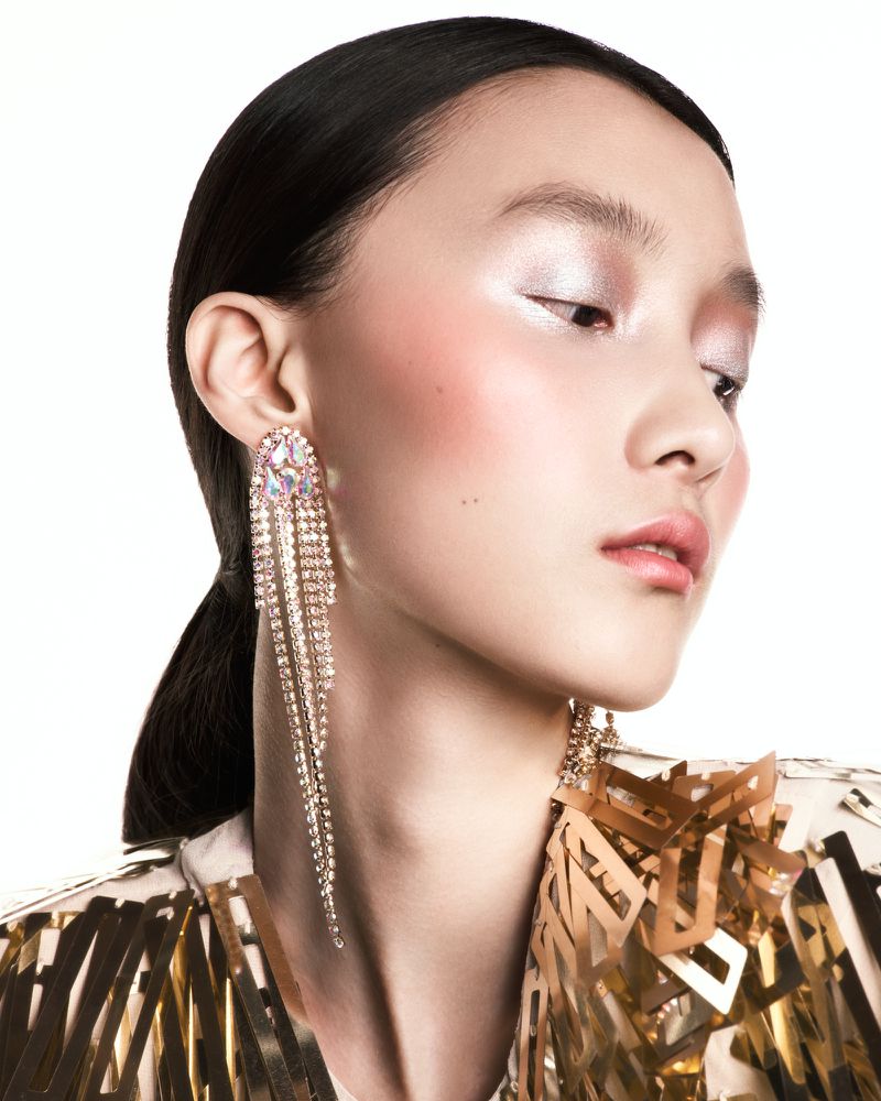 Model Mengyao Wang wearing long rhinestone fringe earrings and a metallic gold geometric top against a pure white background.