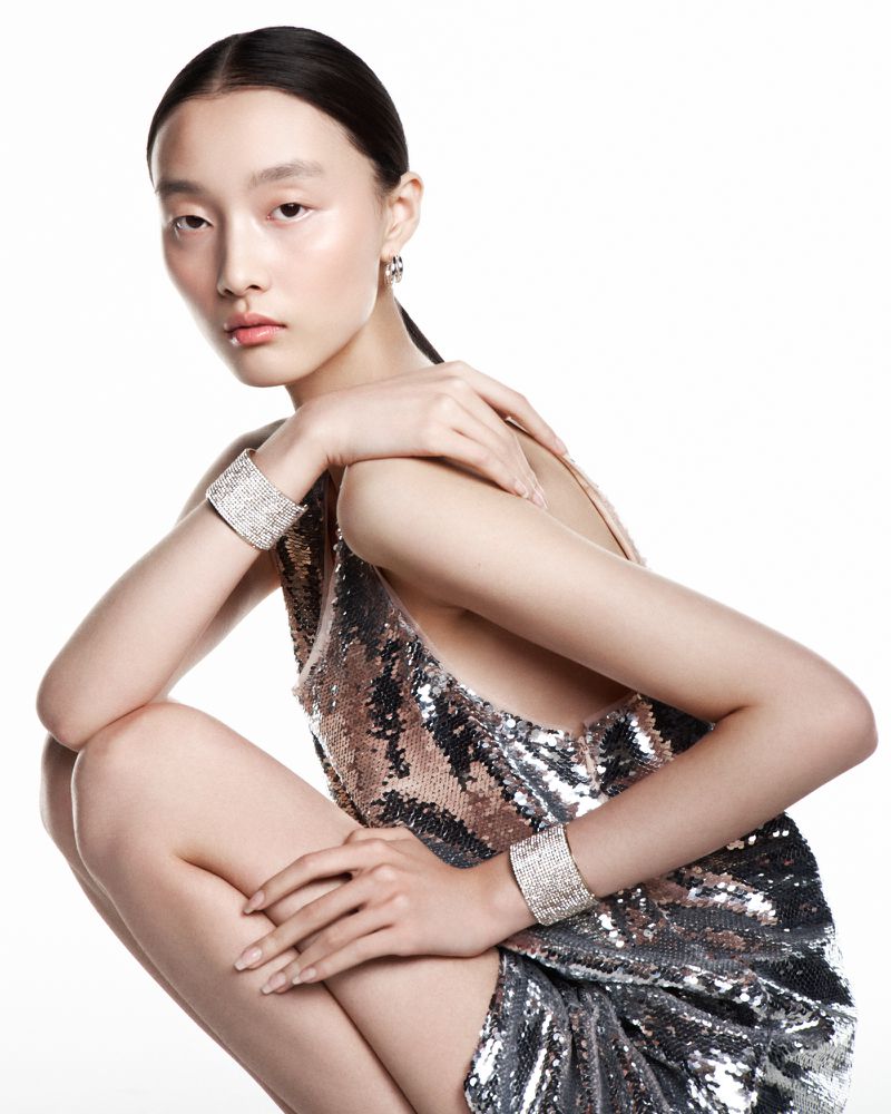 East Asian model Mengyao Wang crouching in a shimmering silver sequin dress and crystal cuff bracelets against a white backdrop.