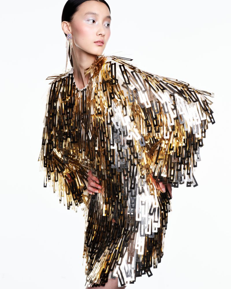 Model Mengyao Wang wearing a metallic gold geometric fringe dress and drop earrings against a white studio background.