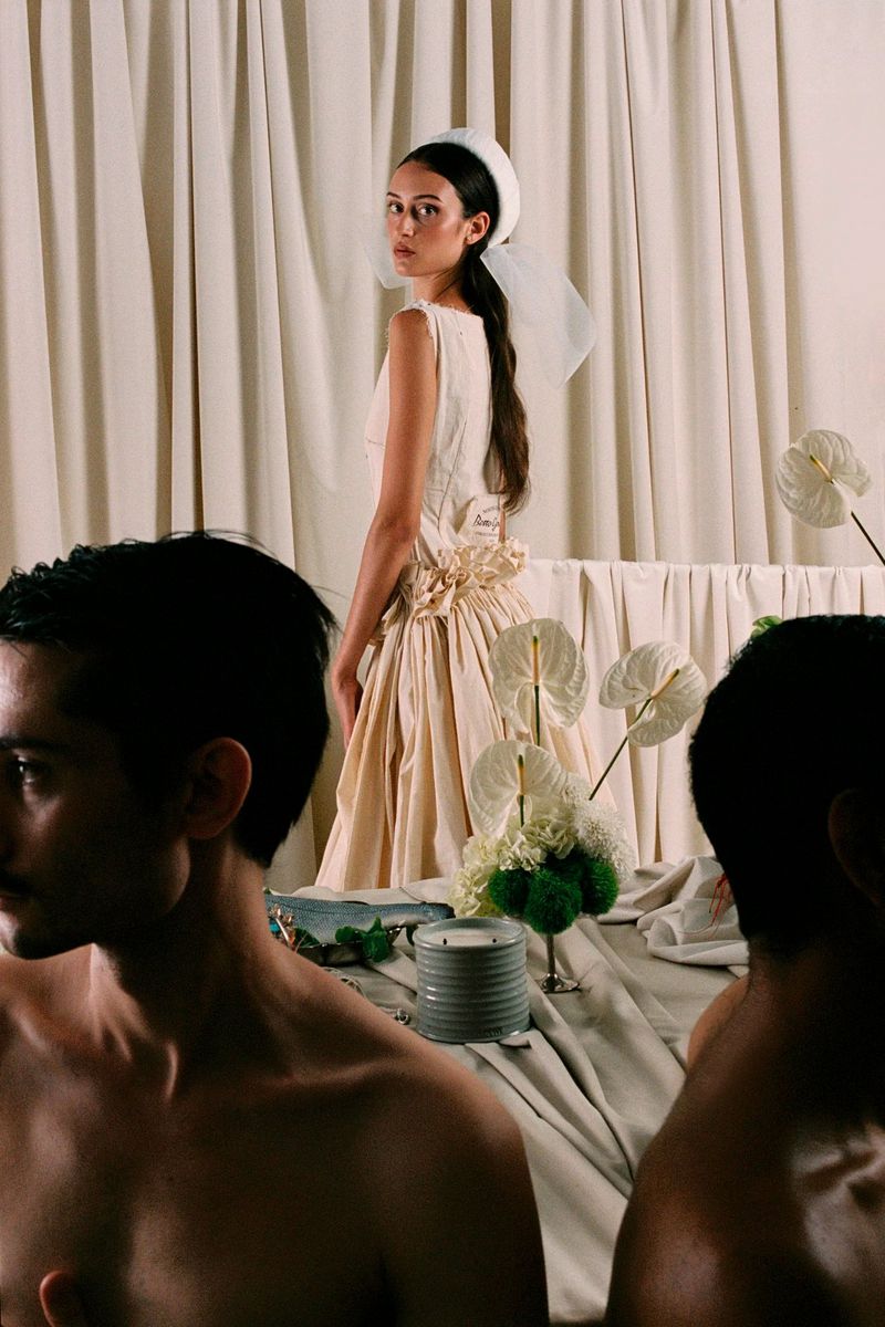 Woman in vintage-style dress posing with floral arrangement on draped table, two bare-chested men sit at the table; indoor setting.