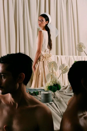 Woman in vintage-style dress posing with floral arrangement on draped table, two bare-chested men sit at the table; indoor setting.