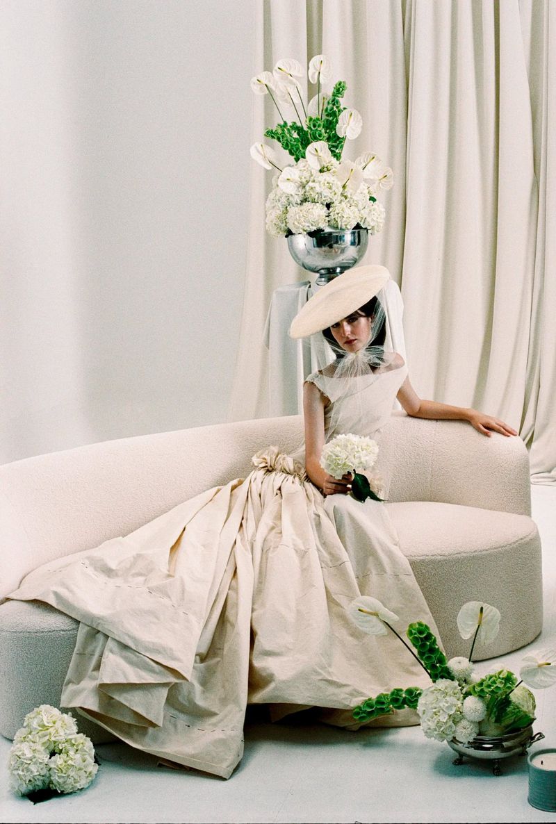 Young woman in a beige gown and hat sitting on a sofa surrounded by white floral arrangements in a light and airy photo studio.
