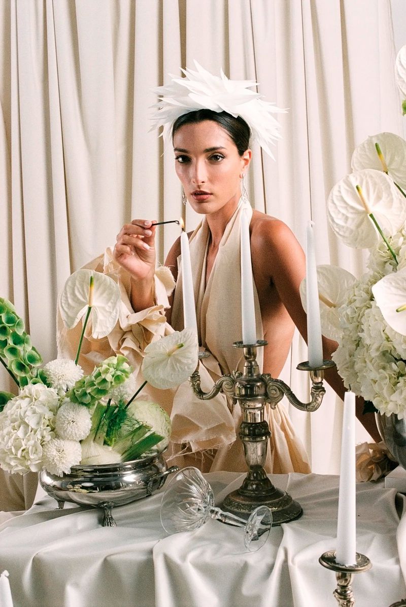 Elegant portrait of a woman lighting candles, wearing a feathered headpiece, surrounded by white flowers against a draped backdrop.
