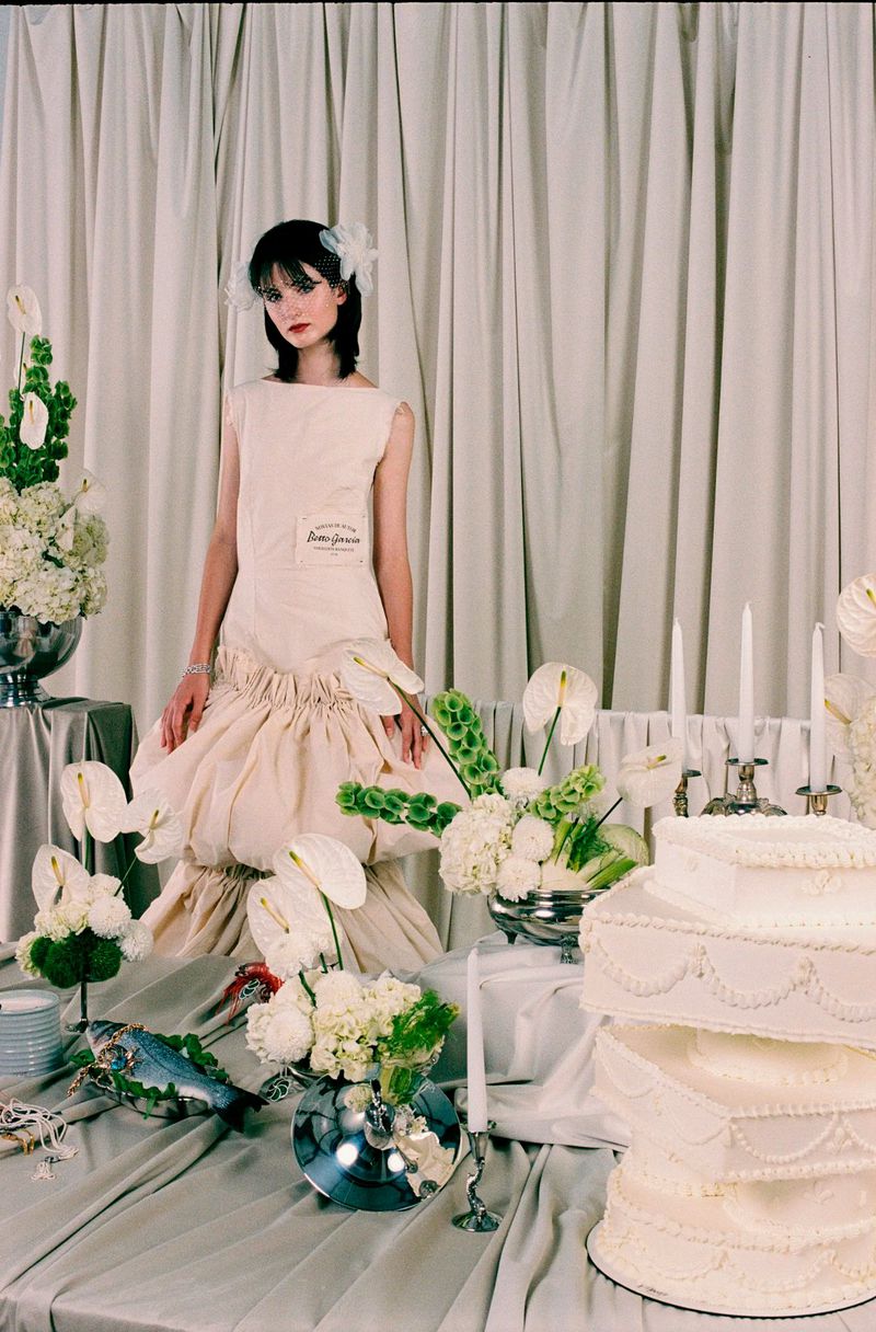 Young woman poses with a neutral expression in a sleeveless, ruffled dress, surrounded by cakes and flowers against a draped backdrop.