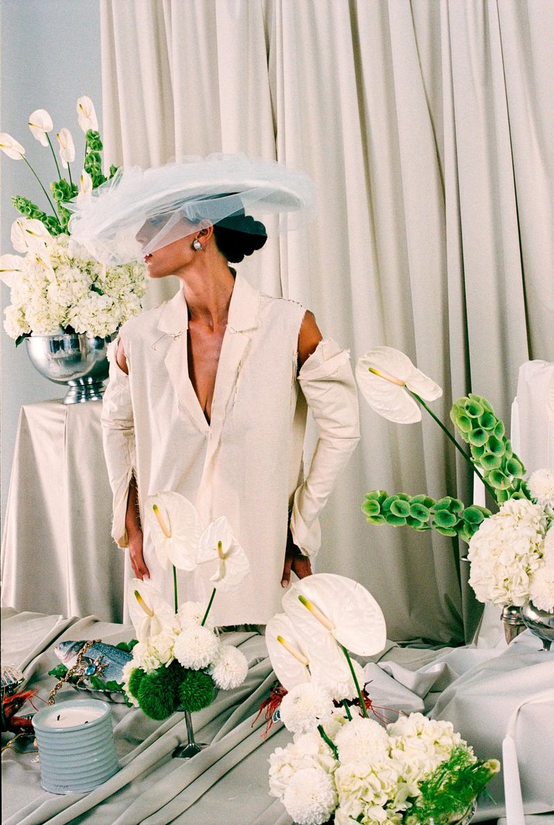 Woman in oversized white jacket with cutouts, wearing a sheer hat, surrounded by white flowers in an indoor studio setting.