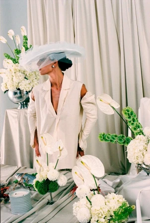 Woman in oversized white jacket with cutouts, wearing a sheer hat, surrounded by white flowers in an indoor studio setting.