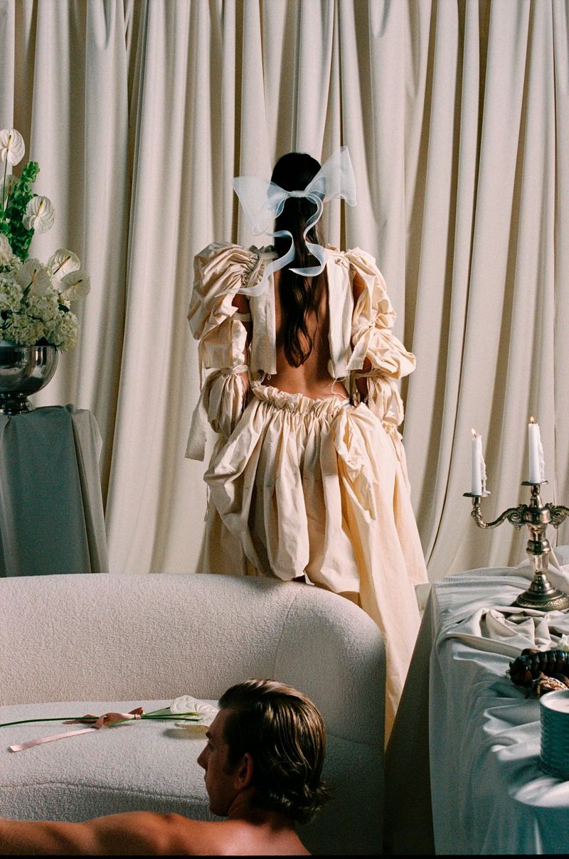 Woman in a cream dress with puffy sleeves stands in front of draped curtains; a man lies on a white couch in the foreground.